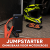 Een compacte zwarte jumpstarter met accuklemmen, klaargelegd op een crossbusvloer naast een helm en handschoenen
