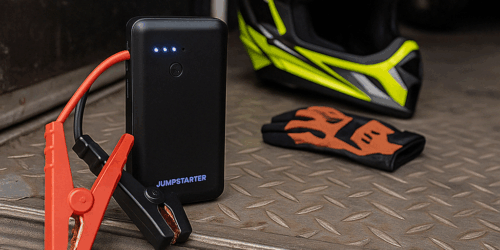 Een compacte zwarte jumpstarter met accuklemmen, klaargelegd op een crossbusvloer naast een helm en handschoenen