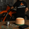 Een werkbank met motoronderdelen waaronder een zuiger, luchtfilter, olie en een jumpstarter, met een motorcrossrijder sleutelend op de achtergrond