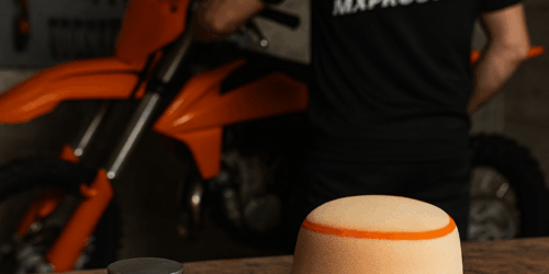 Een werkbank met motoronderdelen waaronder een zuiger, luchtfilter, olie en een jumpstarter, met een motorcrossrijder sleutelend op de achtergrond