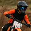 Een motorcrosser met spiegelende crossbril in actie op een modderige baan, modderspatten vliegen omhoog richting het gezicht