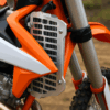 Een zilveren radiator guard op een witte en oranje crossmotor, gemonteerd met bouten en zichtbaar tussen de kappen