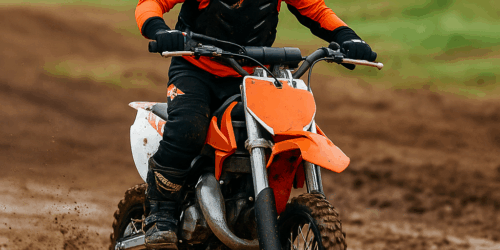 Een jonge rijder op een semi-automatische crossmotor rijdt over een modderige baan, met helm, bril en beschermende kleding in fel oranje en zwart